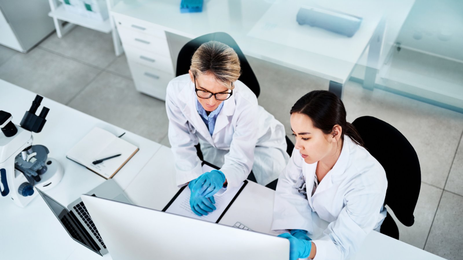 High-angle view of two scientists in white lab coats and blue gloves reviewing data on a computer monitor.